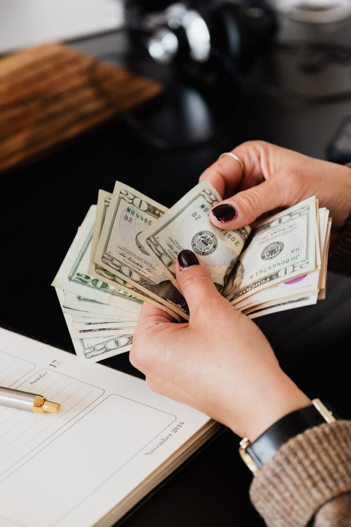 Unrecognizable elegant female in sweater counting dollar bills while sitting at wooden table with planner and pen
