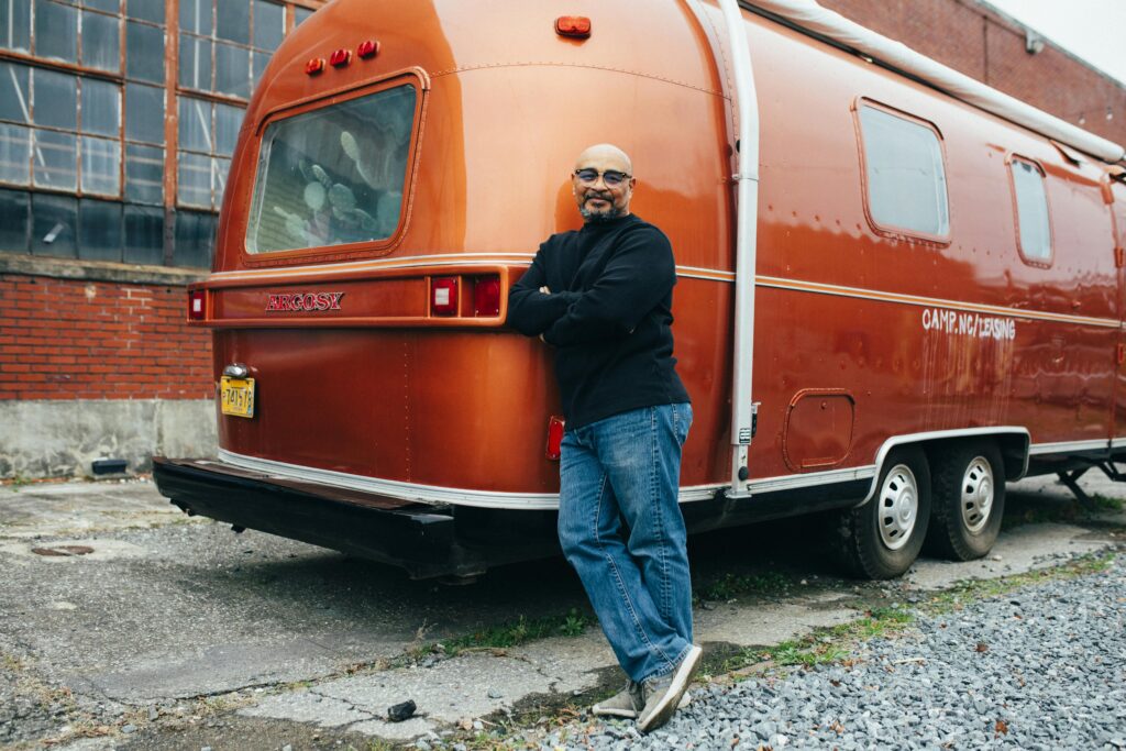 A man stands confidently against a vintage orange camper van in an industrial setting.