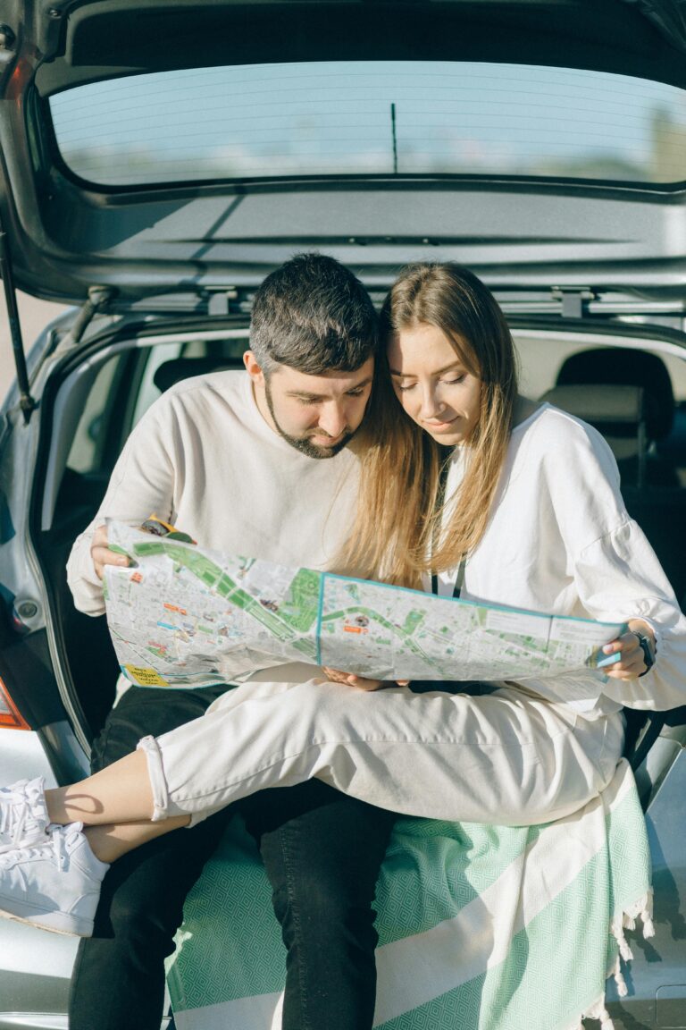 A couple sitting in a car trunk studying a map during a sunny road trip.